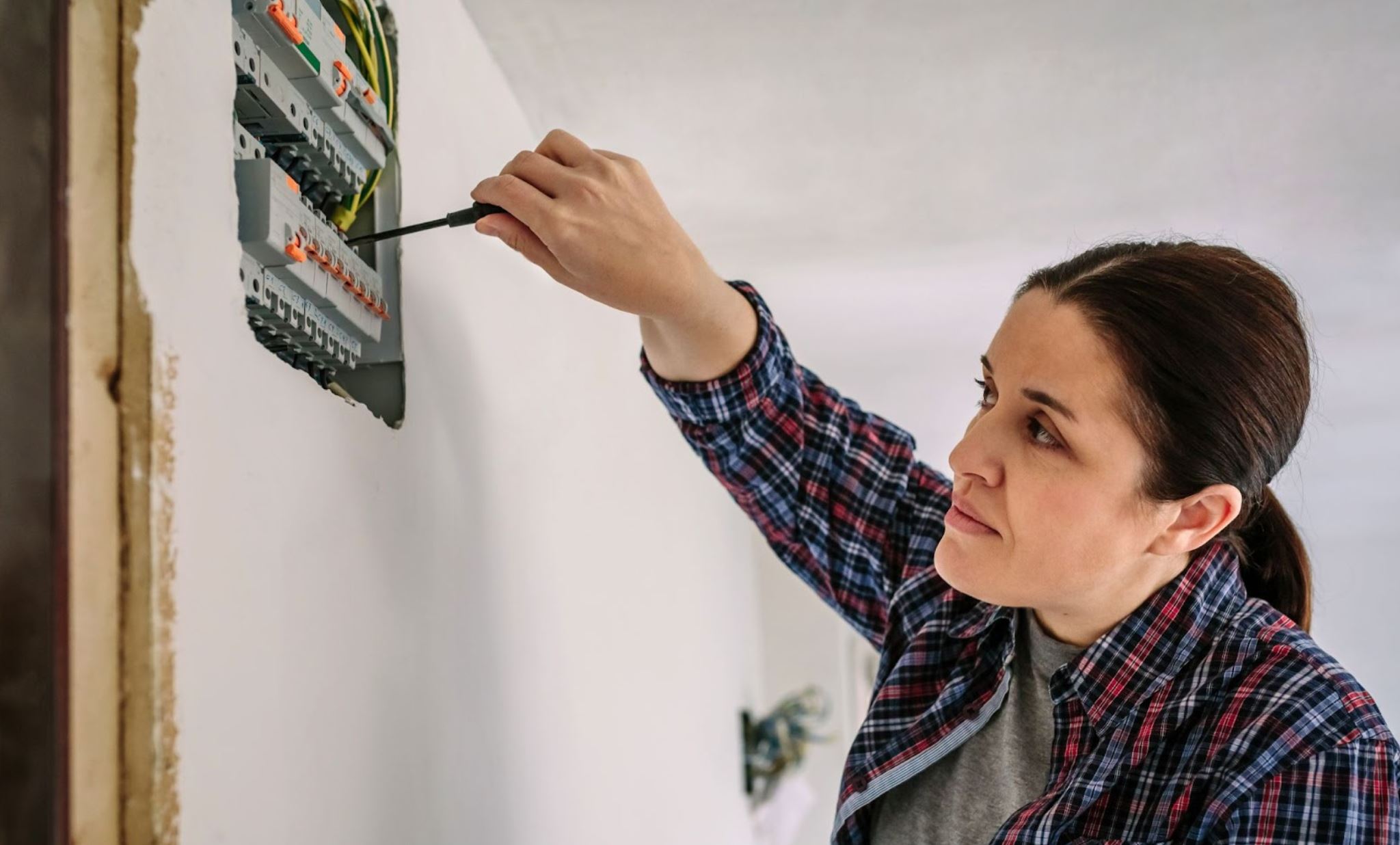 A female electrician in a checkered, long-sleeved polo fixes a home's distribution panel with a screwdriver.