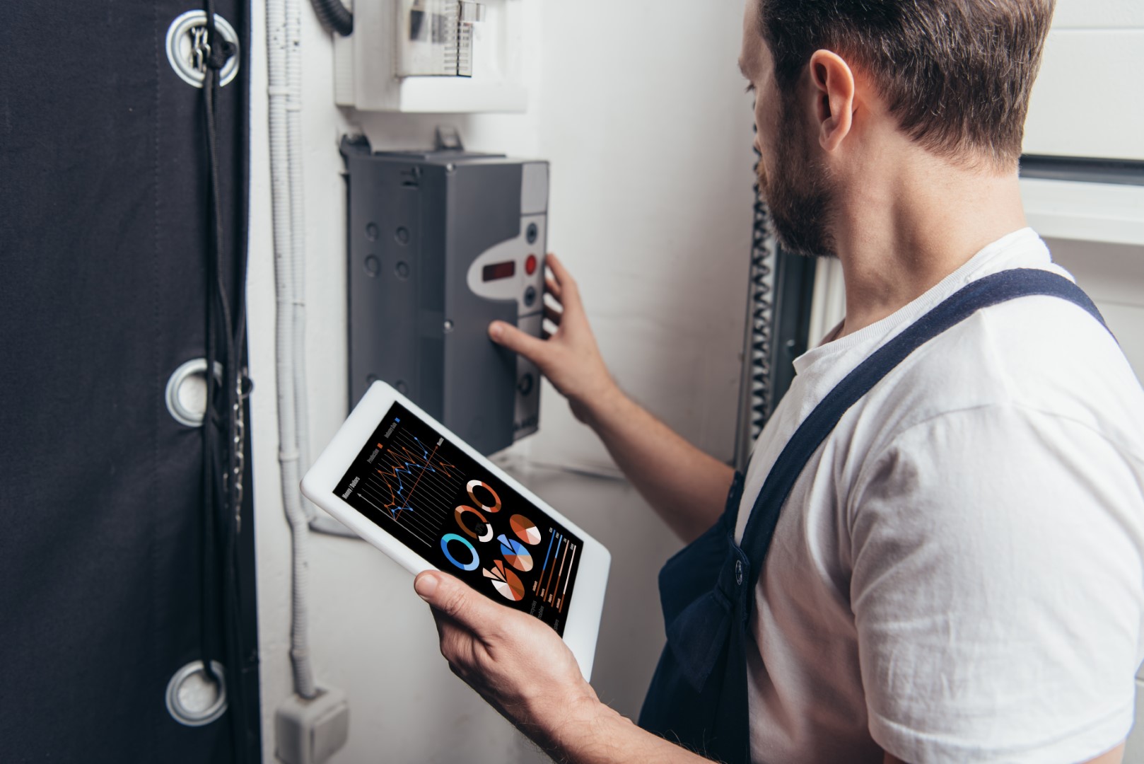 A male electrician holding a digital tablet with graphs on the screen and checking an electric breaker box.