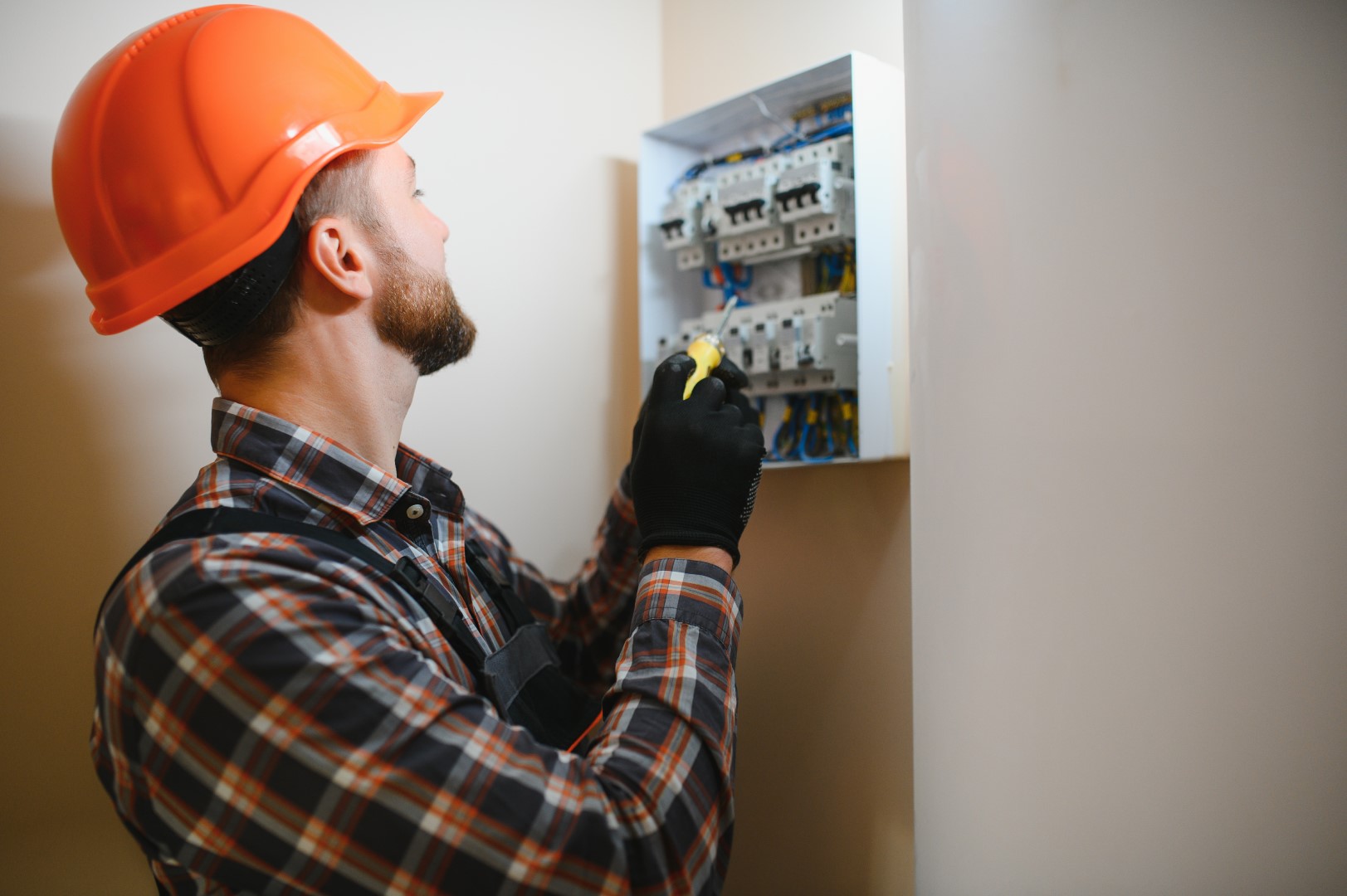 A male electrician wearing an orange hard hat, a plaid shirt, and black gloves is working on a switchboard with an electrical connecting cable.