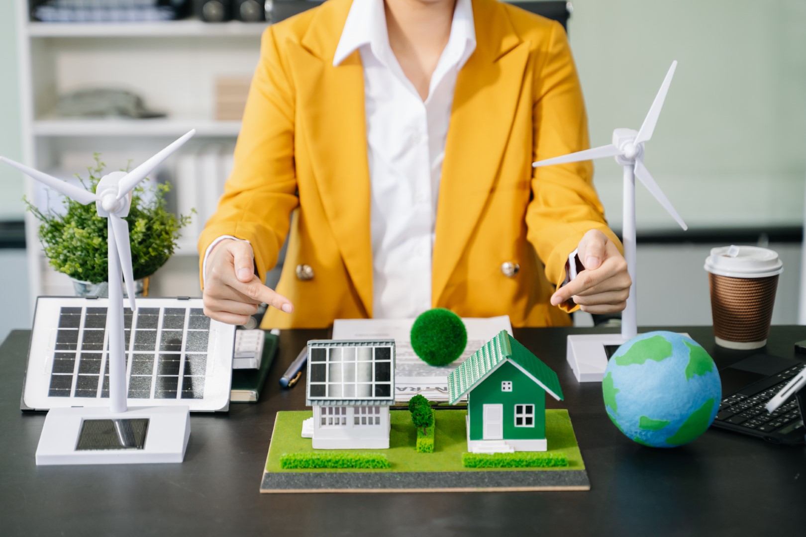 A miniature example of a house with solar panels next to windmills and a globe on a desk with a woman wearing yellow points at it.