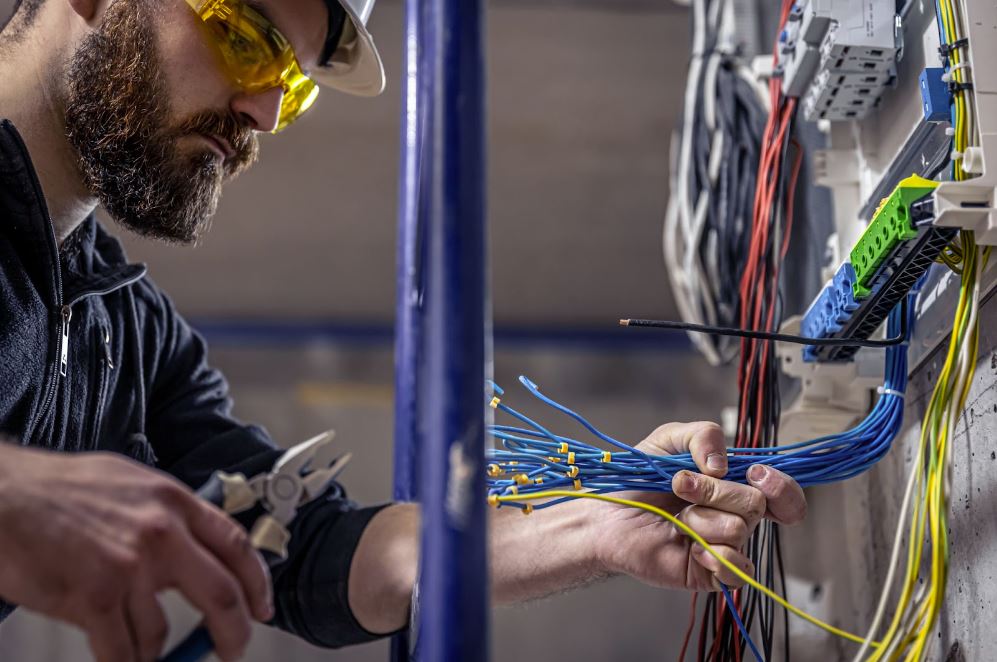 A male electrician wearing a helmet and safety goggles attaches color-coded wires to a switchboard, focusing carefully on proper connections.