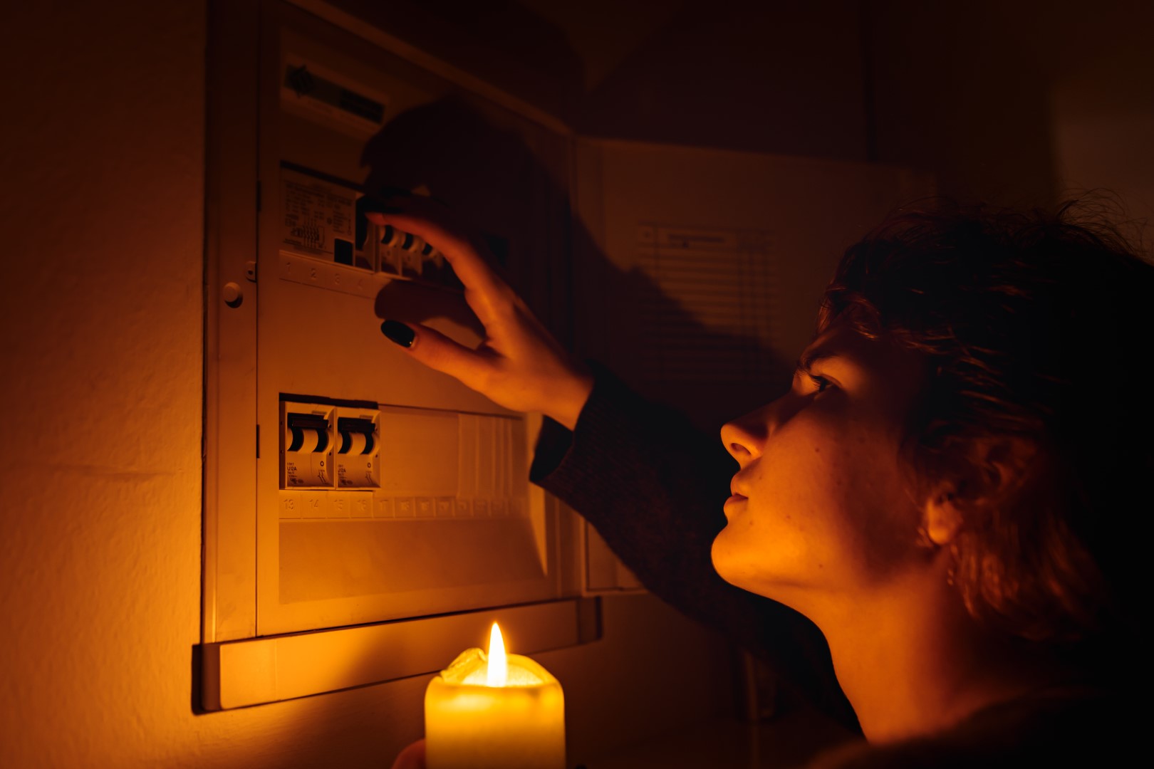 A woman holding a candle in darkness examines a fuse box or electrical distribution board at home during a power outage.