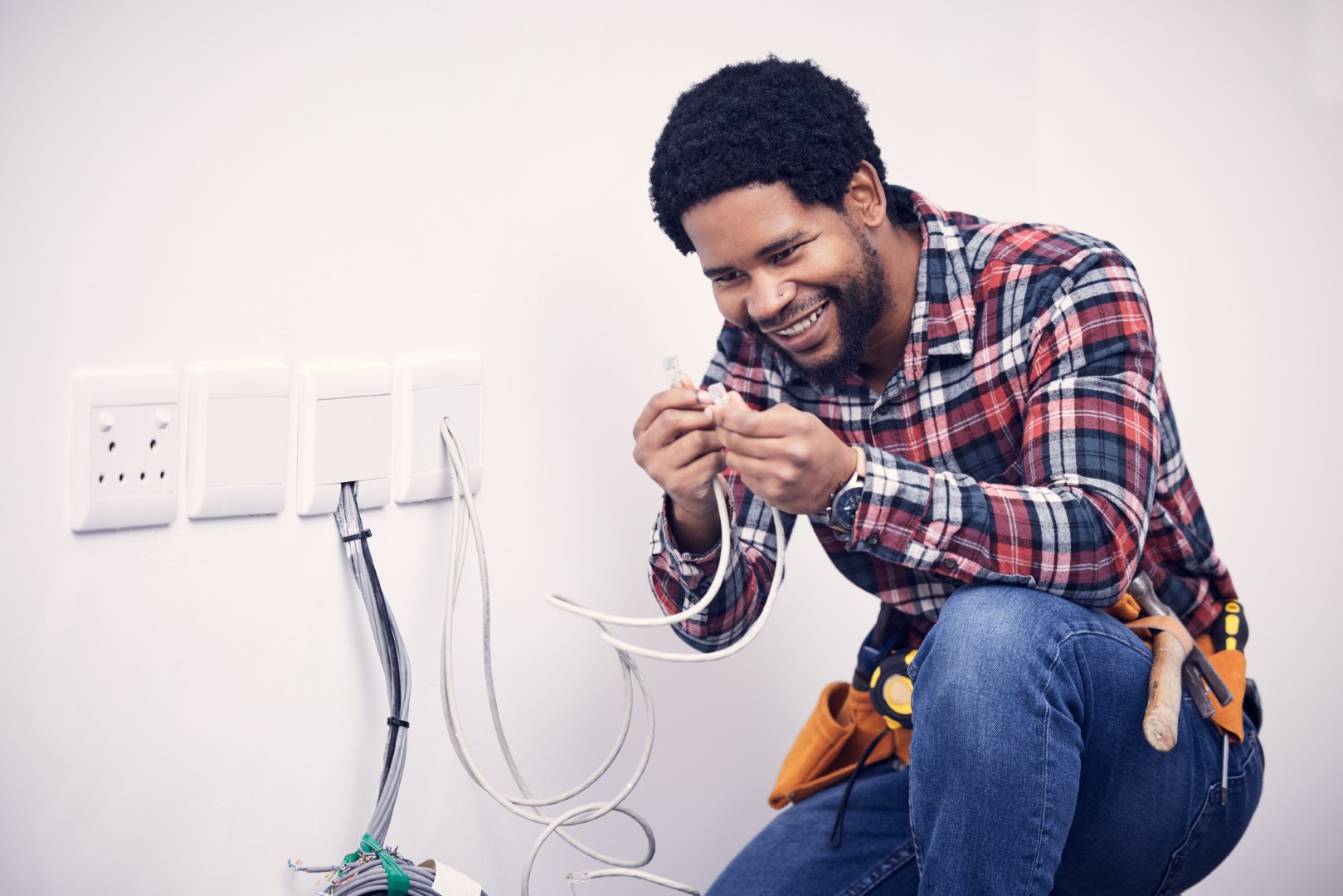 An electrician wearing a plaid shirt, jeans, and a utility belt works on a home switchboard.