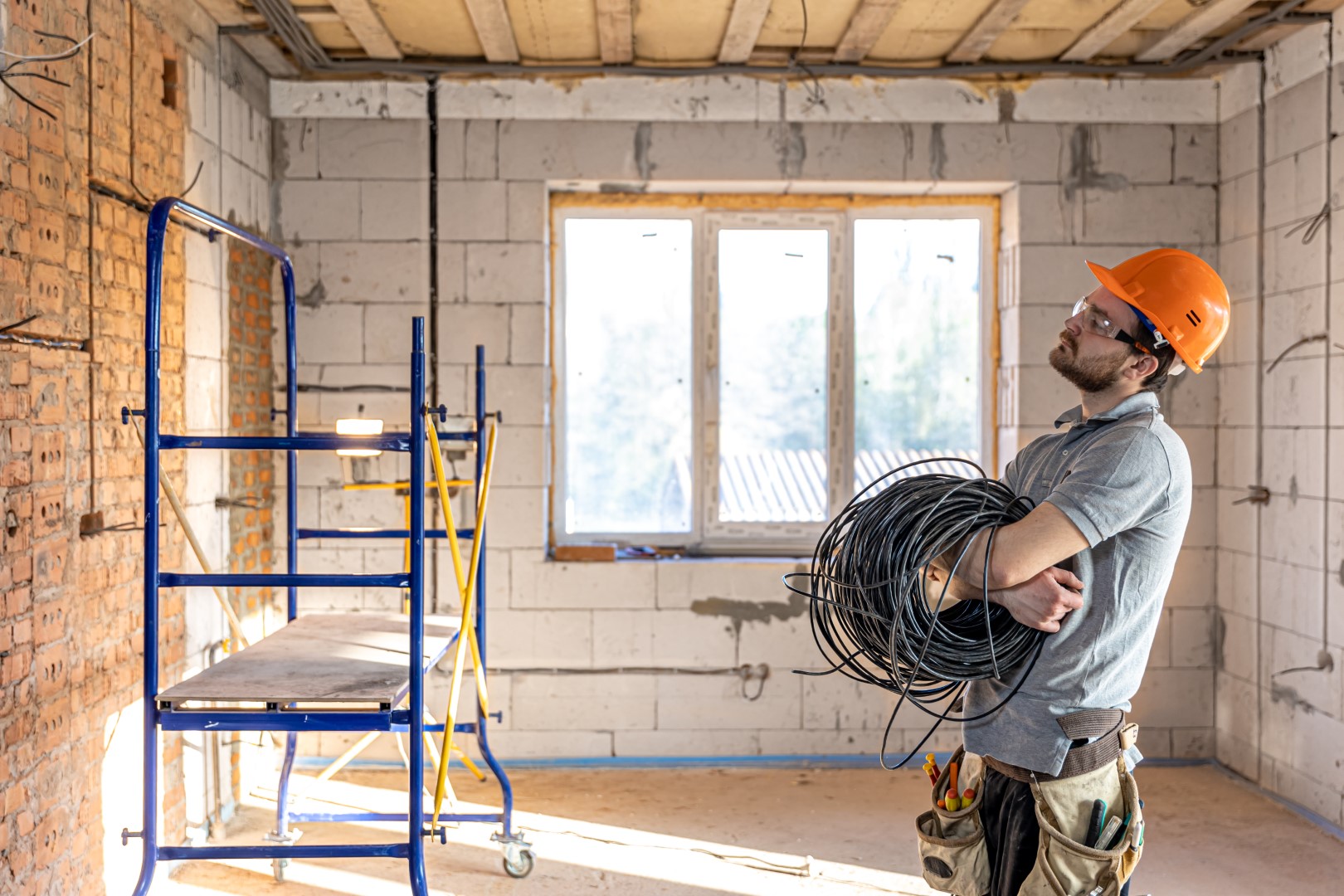 An electrician wearing an orange hard and a tool belt holding rolled-up electrical cables while looking at a wall at a construction site.