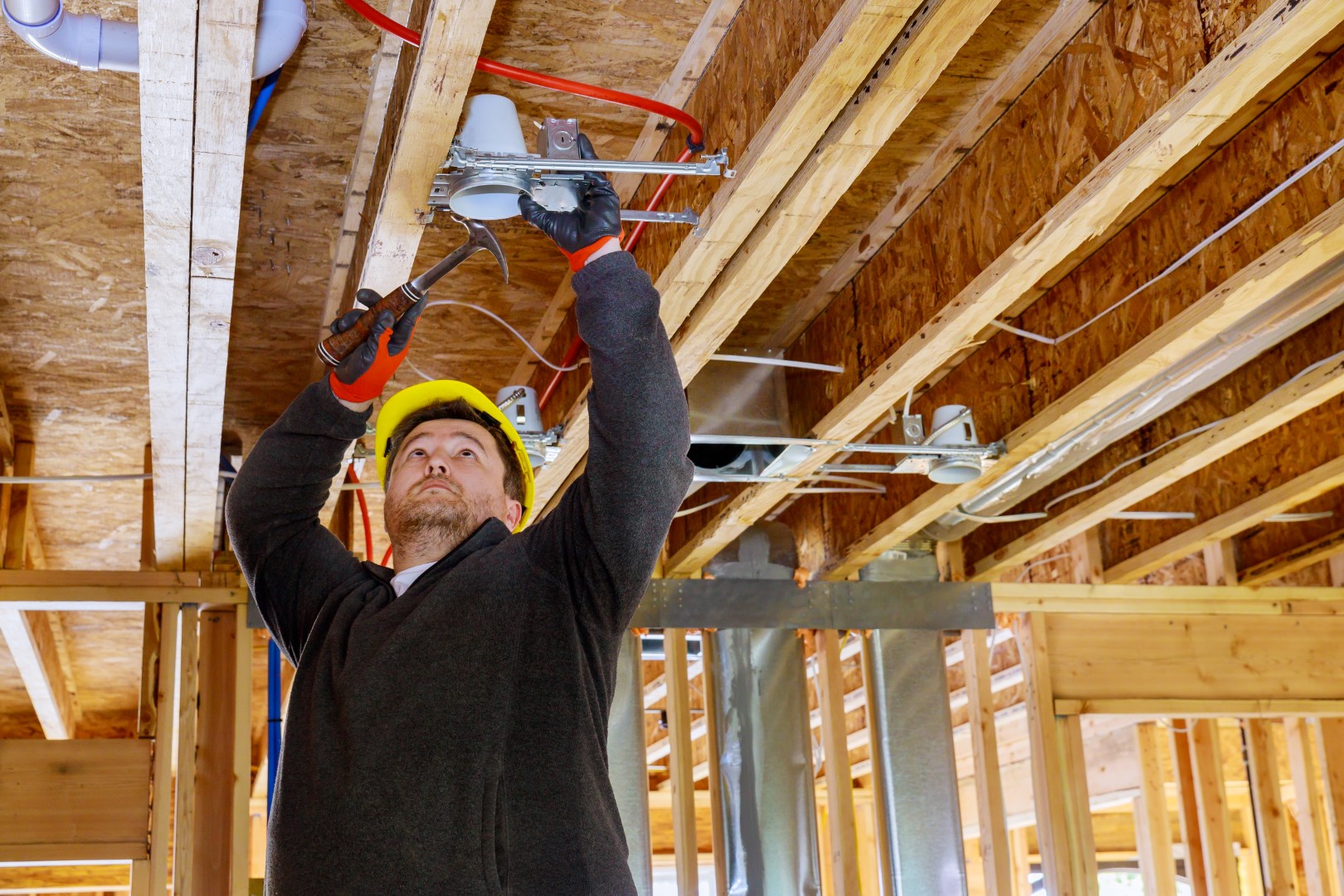 An electrician wearing gloves, a jacket, and a hard hat installs electrical wiring for a ceiling light.