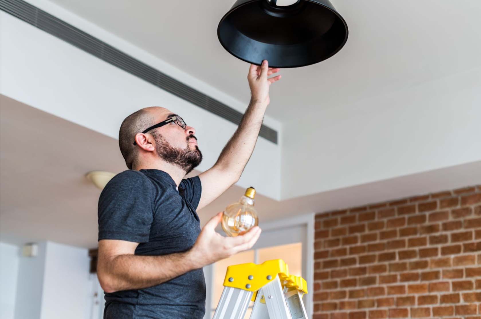 A balding man with a well-shaven beard, wearing a black shirt, stands on an aluminum ladder holding a light bulb, about to install it in a black hanging lamp, demonstrating household electrical repairs.