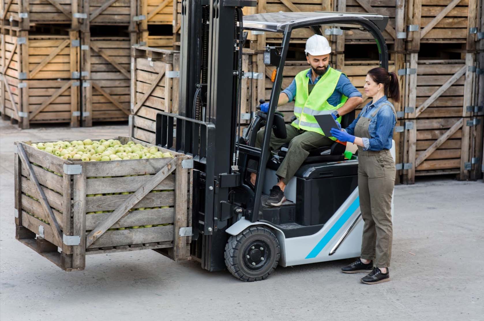 A bearded forklift driver wearing safety gear discusses loading logistics with the female warehouse manager in brown overalls as a crate of fruits sits ready on the forklift loader; stacks of crates piled in steel shelves are visible in the background.