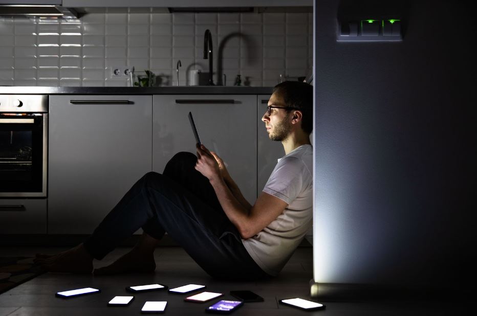 A bearded man wearing black-framed eyeglasses, a white shirt, and black pants uses a tablet to chat on social media during a blackout. 