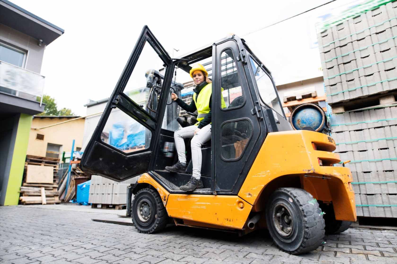 A female forklift truck driver in a safety uniform alights from a black and yellow forklift at an outdoor warehouse, with piles of gray crates stacked behind her. On her left is a gray and green building.