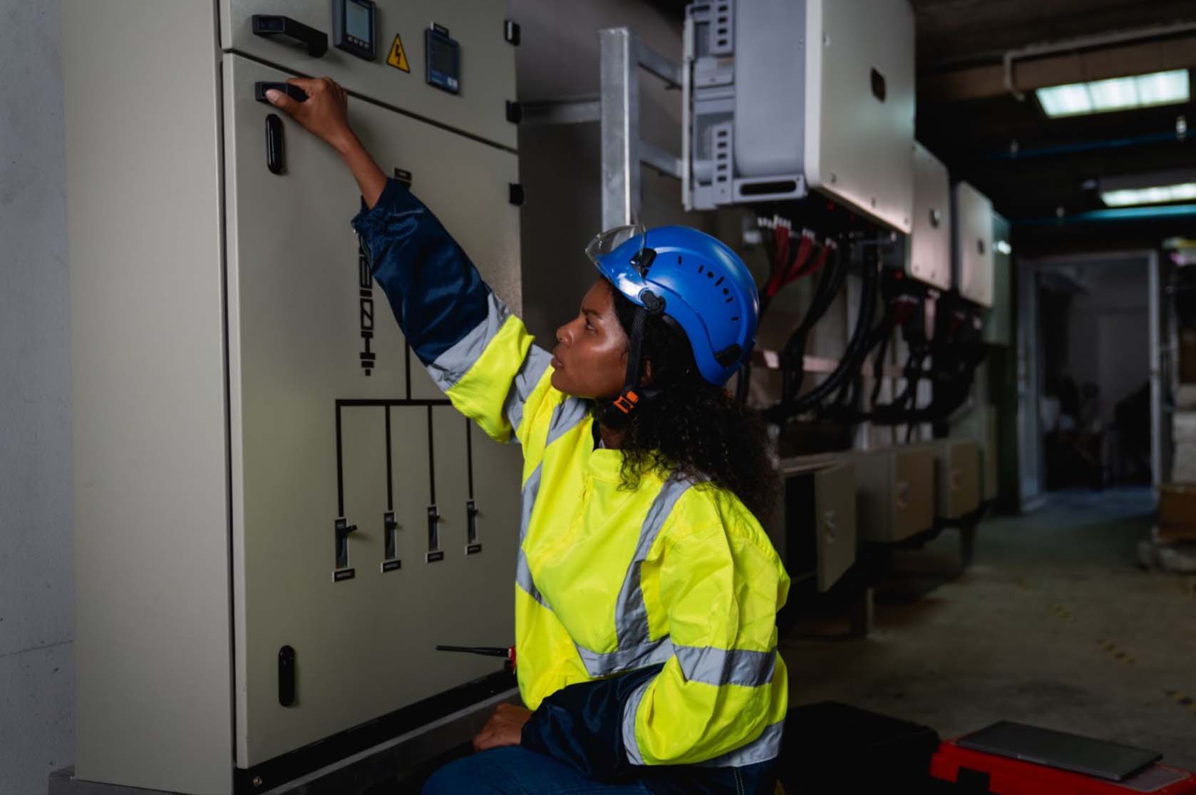 A female licensed electrician wearing safety gear inspects an electrical switchboard, verifying the operational voltage range in a control room.