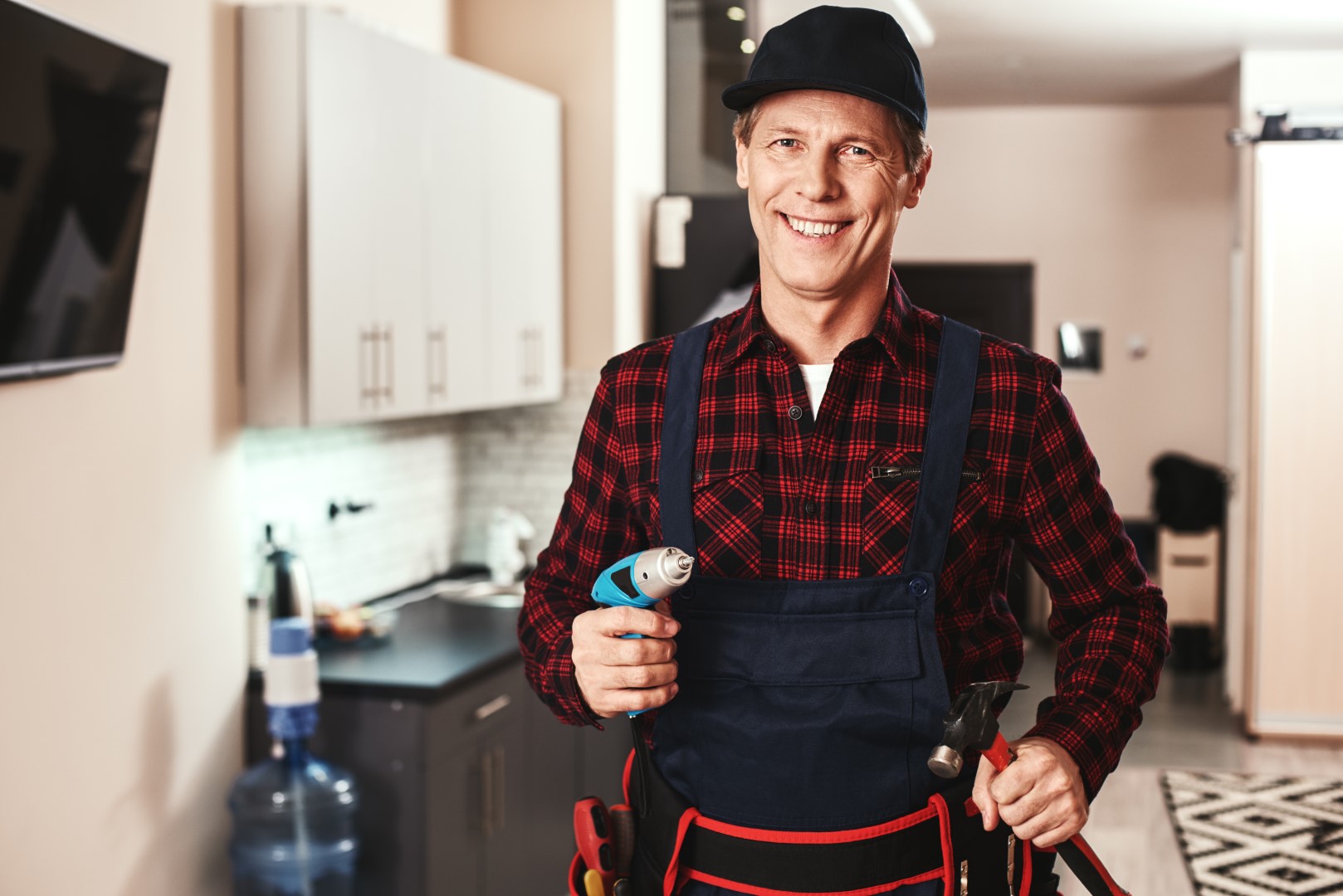 A home repairs professional wearing a red plaid shirt, overalls, a tool belt, and a black hat holding a drill while smiling in a kitchen.