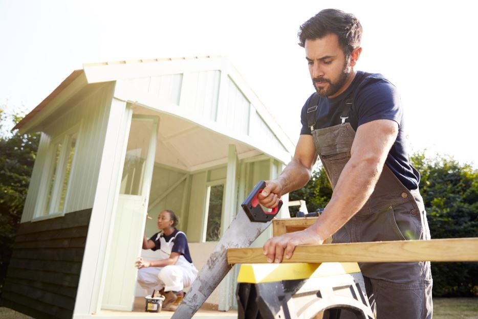 A male carpenter and a female apprentice sawing wood together to build an outdoor summerhouse in a garden.