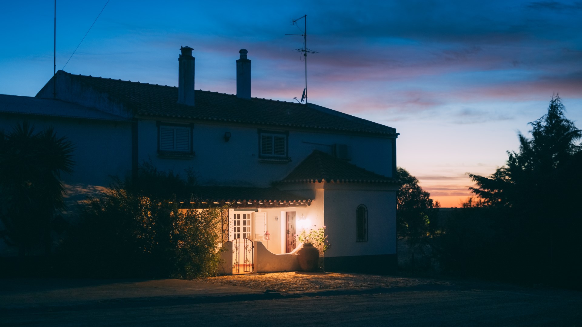 A two-story home surrounded by a few trees in the evening while the sun sets with a porch light on.