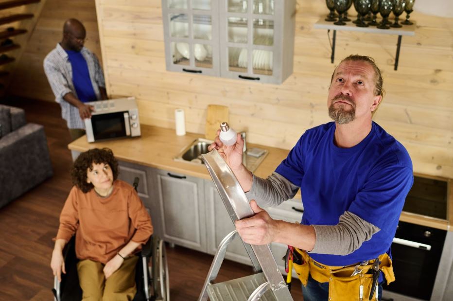 A bearded Caucasian electrician on a stepladder, holding a light bulb and looking upwards, prepares to install it while a woman in a wheelchair watches. 