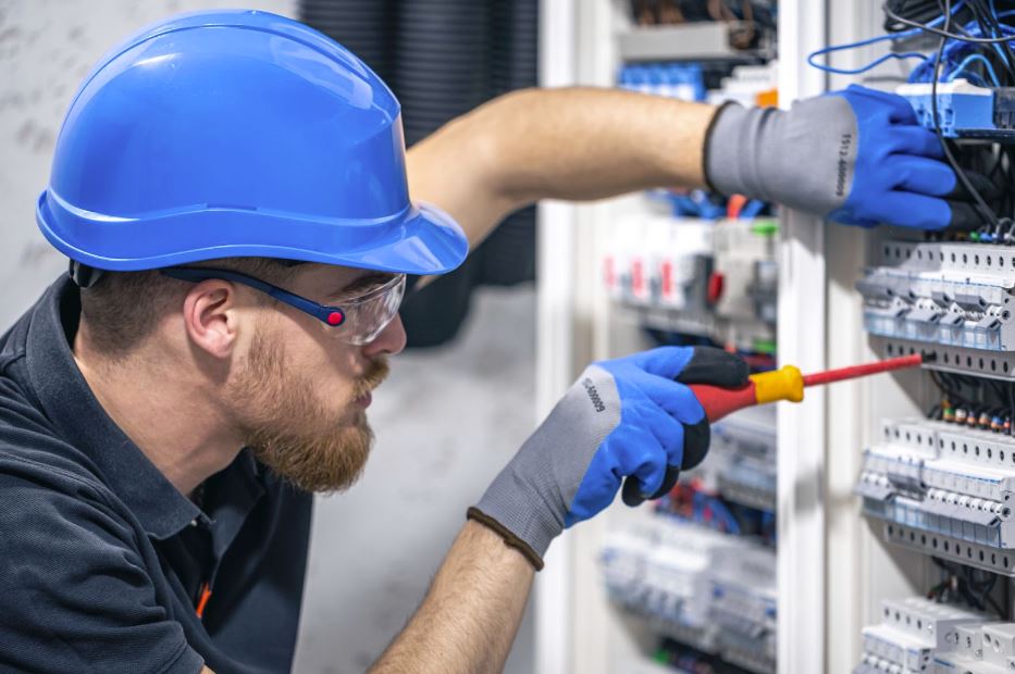 A male electrician wearing a blue helmet, blue and gray gloves, a black shirt, and safety glasses works in a switchboard with fuses, using an orange screwdriver.