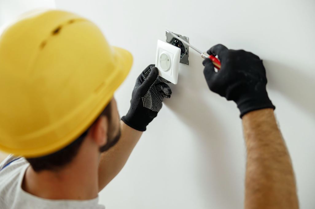 A picture from behind of an electrician wearing a yellow hardhat and gloves using a screwdriver while installing an electrical socket outlet after renovation work.