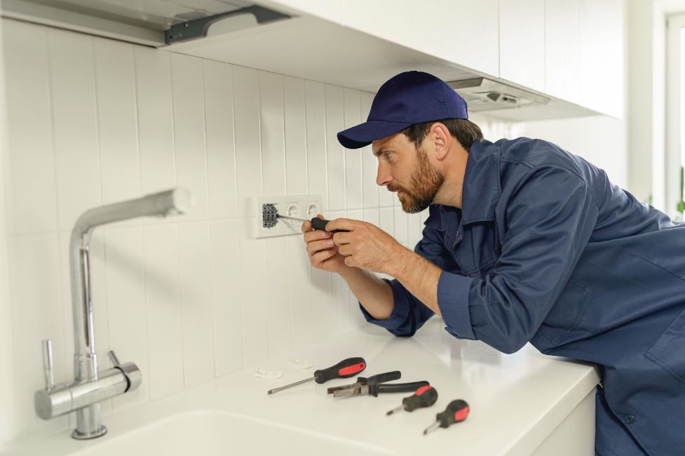 A professional electrician wearing a blue hat and uniform uses a screwdriver while installing a new electrical socket in the home kitchen.