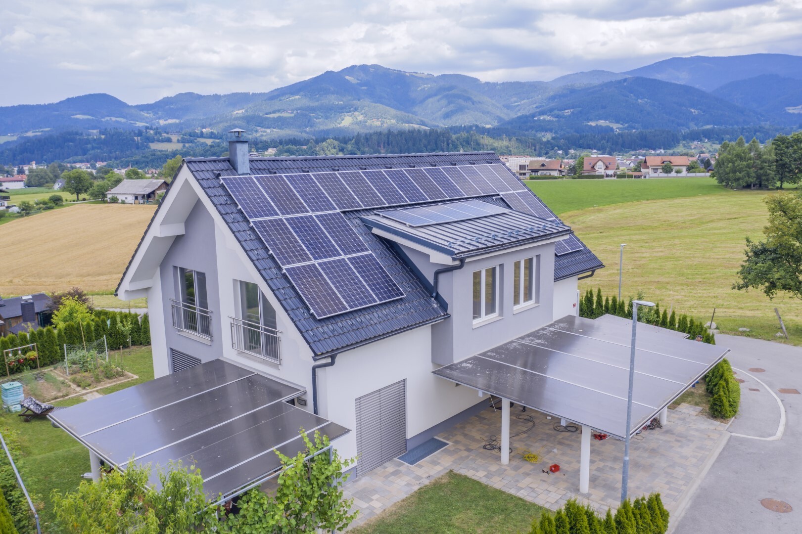 Aerial view of a two-story house with a field and mountains behind it, as well as solar panels on the roof and over the patio area.