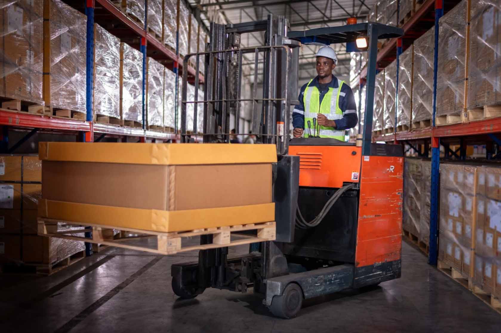 An African male worker in a factory wears a safety uniform while driving a forklift truck, efficiently moving goods and boxes in an industrial setting.