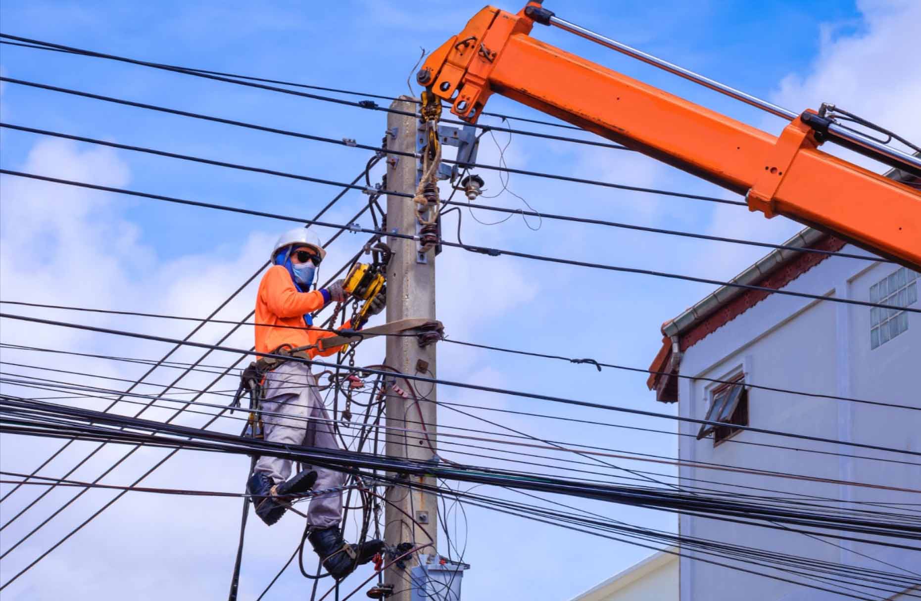 An electrician in a gray hard hat, protective mask, orange long-sleeved shirt, and jeans stands on a crane truck while installing electrical transmission on a power pole, showcasing the work of an electrical provider.