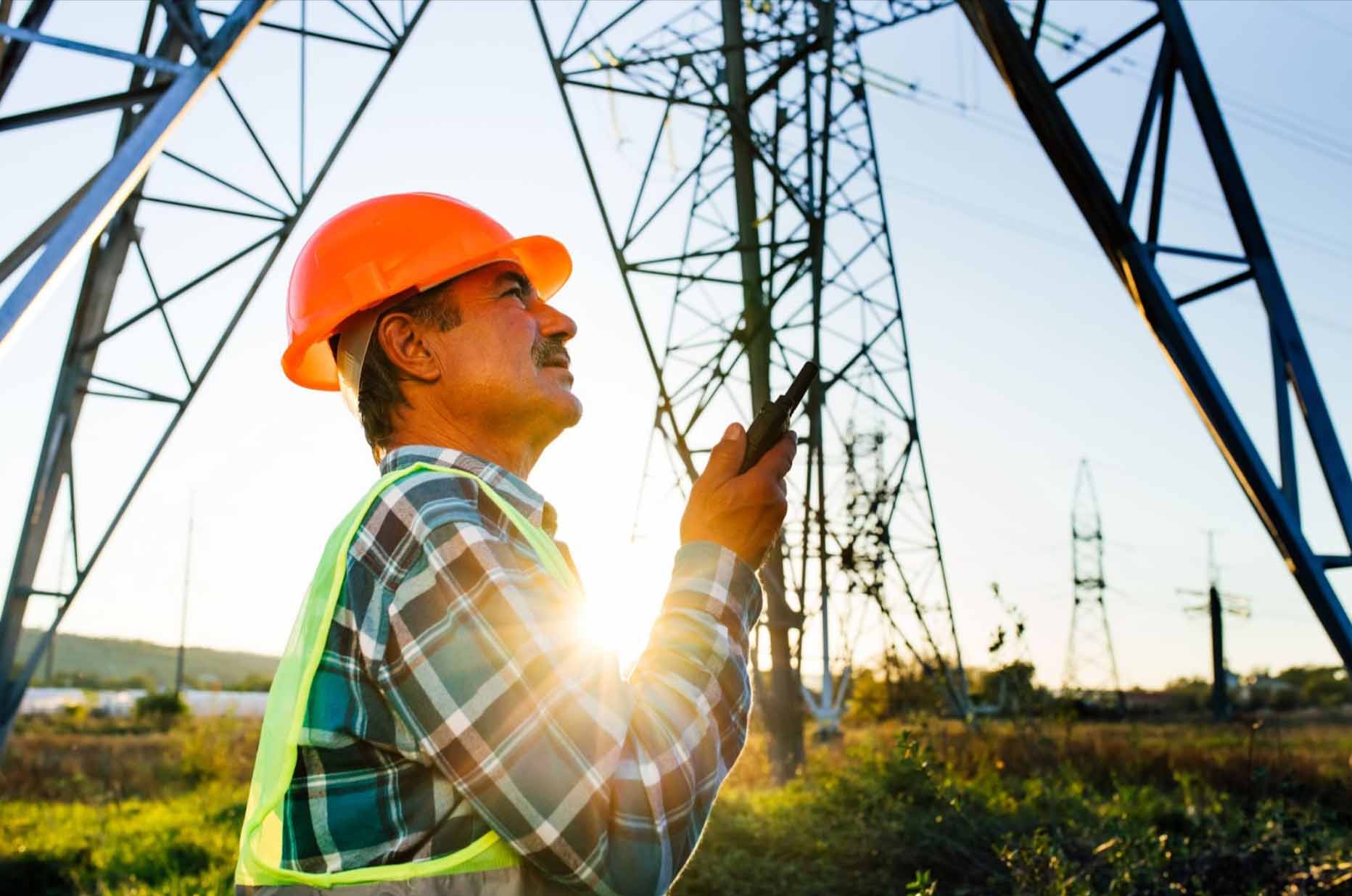 An electrician in an orange hard hat, neon green vest, and checkered long-sleeved polo talking on a portable radio device near a high voltage tower, highlighting electrical provider safety practices.