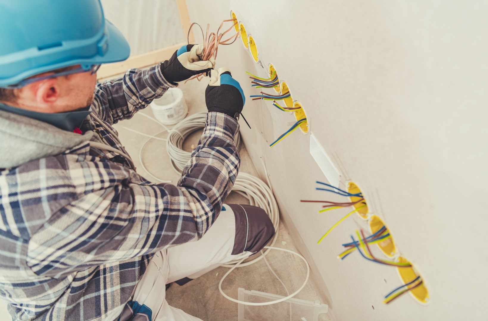 An electrician wearing a plaid hooded jacket and a blue hard hat is installing wiring for electrical outlets.