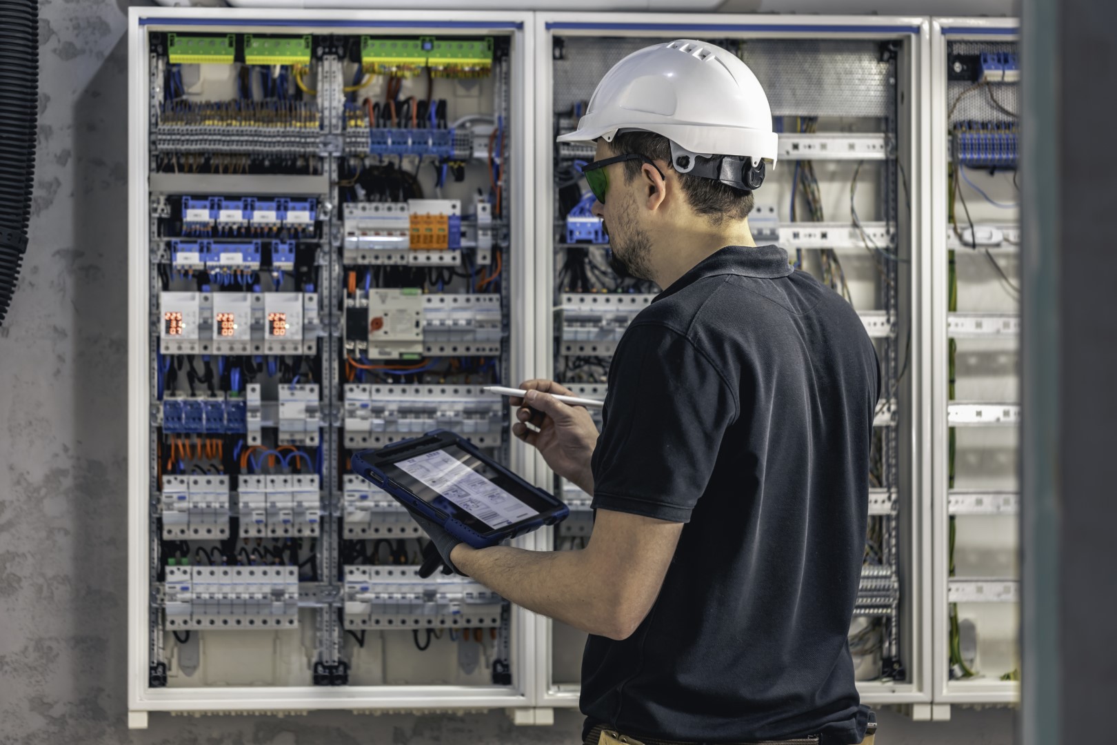 An electrician wearing a white hard hat and a black shirt using a smart tablet to install and connect electrical equipment.