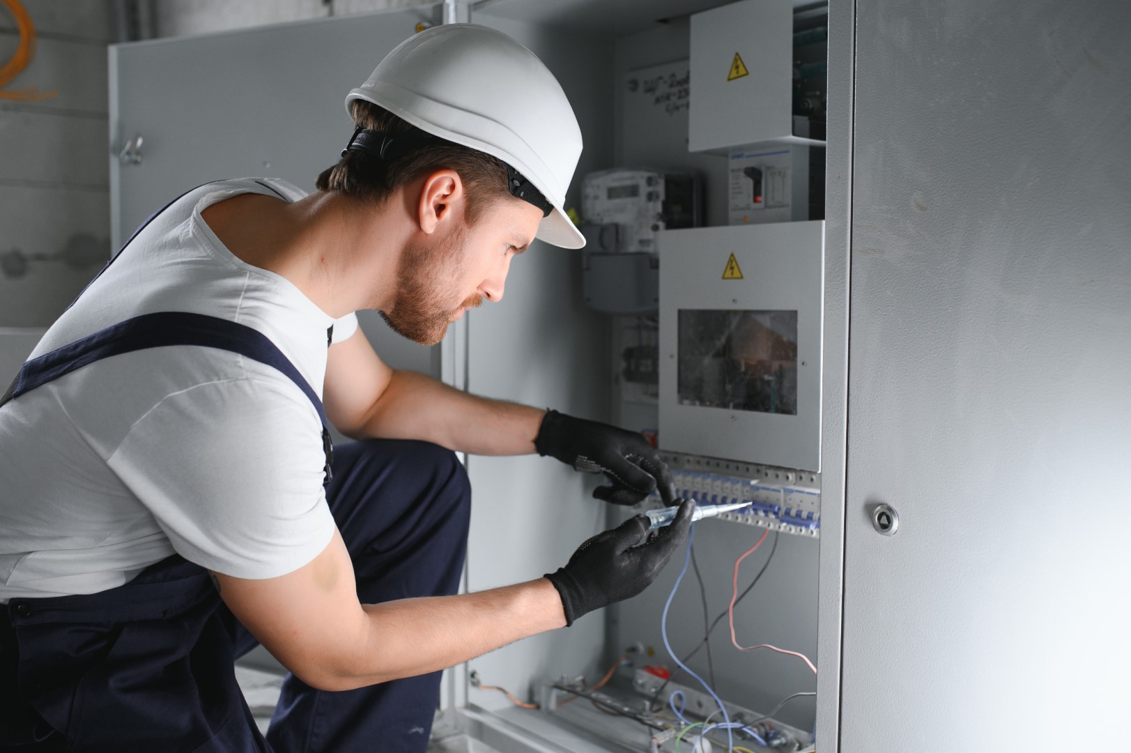 An electrician wearing a white hard hat and overalls using a tool to fix a circuit breaker.