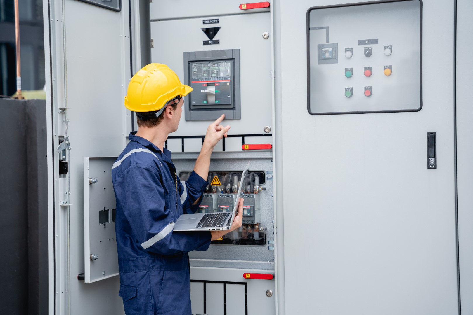 An electrician wearing a yellow hard hat using a laptop inspects the electrical switchboard and verifies the operational voltage range.