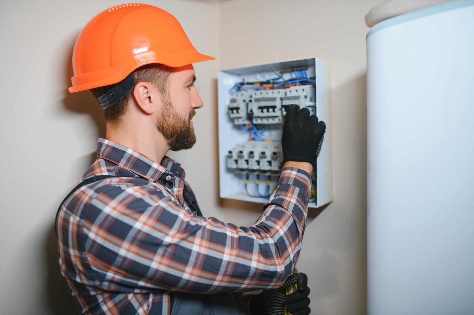 An electrician wearing an orange hard hat and a plaid shirt working on a circuit breaker.