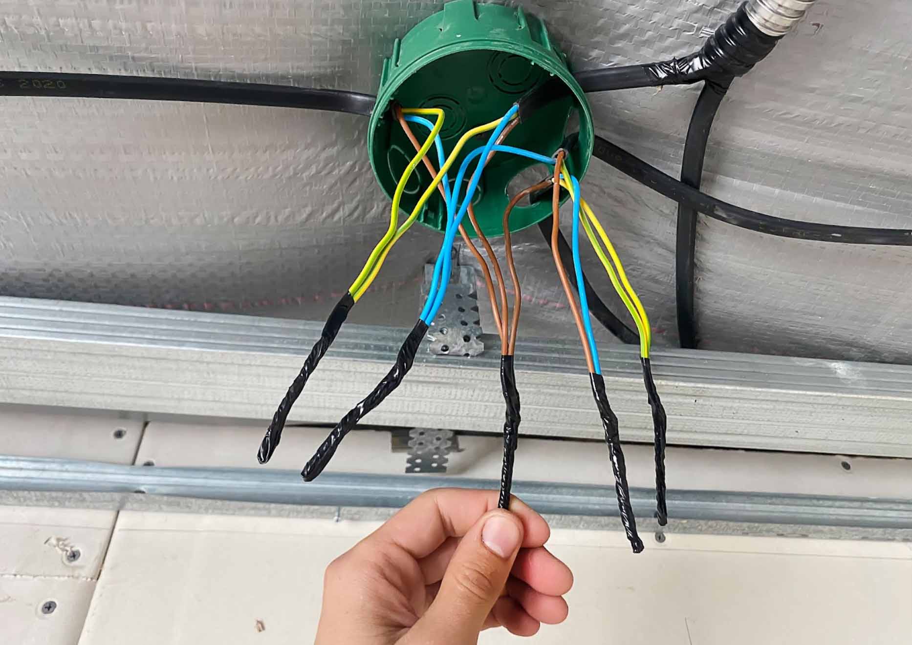 An electrician's hand twisting differently colored wires inside a newly installed green junction box in the ceiling. The insulation mat is shown inside the ceiling.