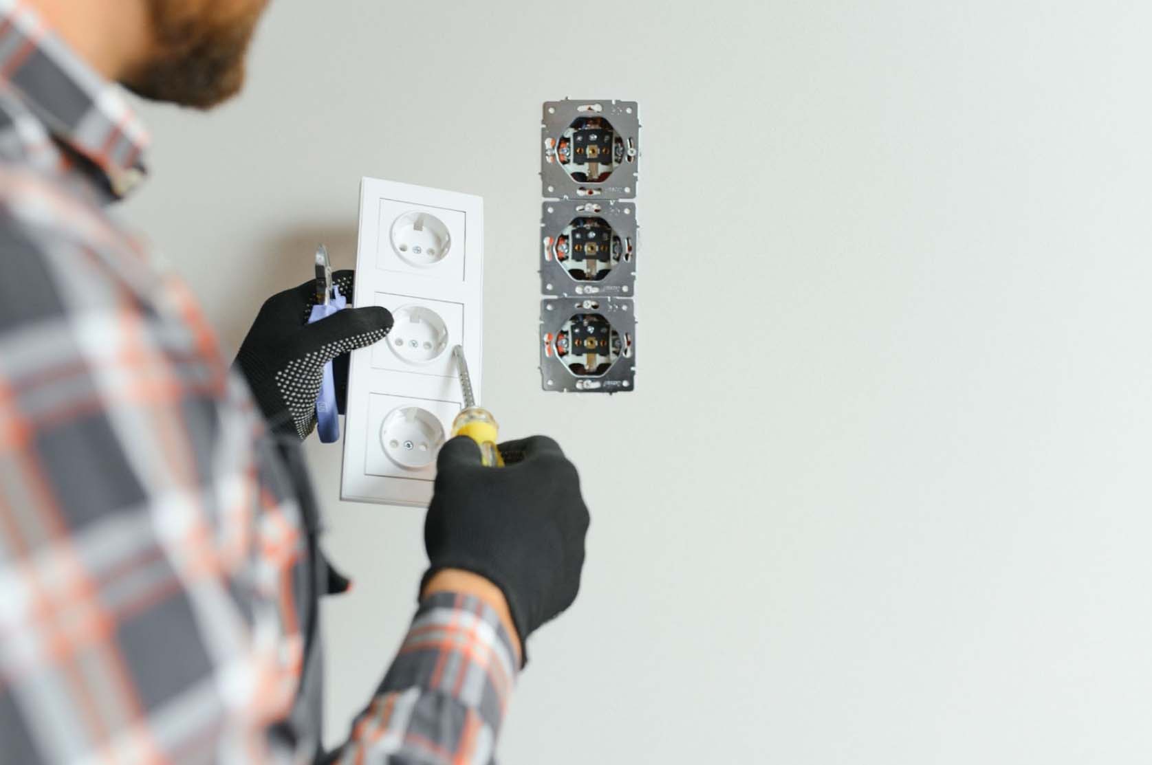 An unidentified bearded man in a checkered long-sleeve polo and black grip gloves uses a screwdriver to repair a power socket in a room.