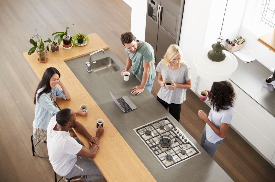 Overhead view of five multicultural friends drinking coffee in a modern kitchen featuring a stove and a sink on a gray granite-topped island, a hanging lamp, and some succulent plants on the countertop.