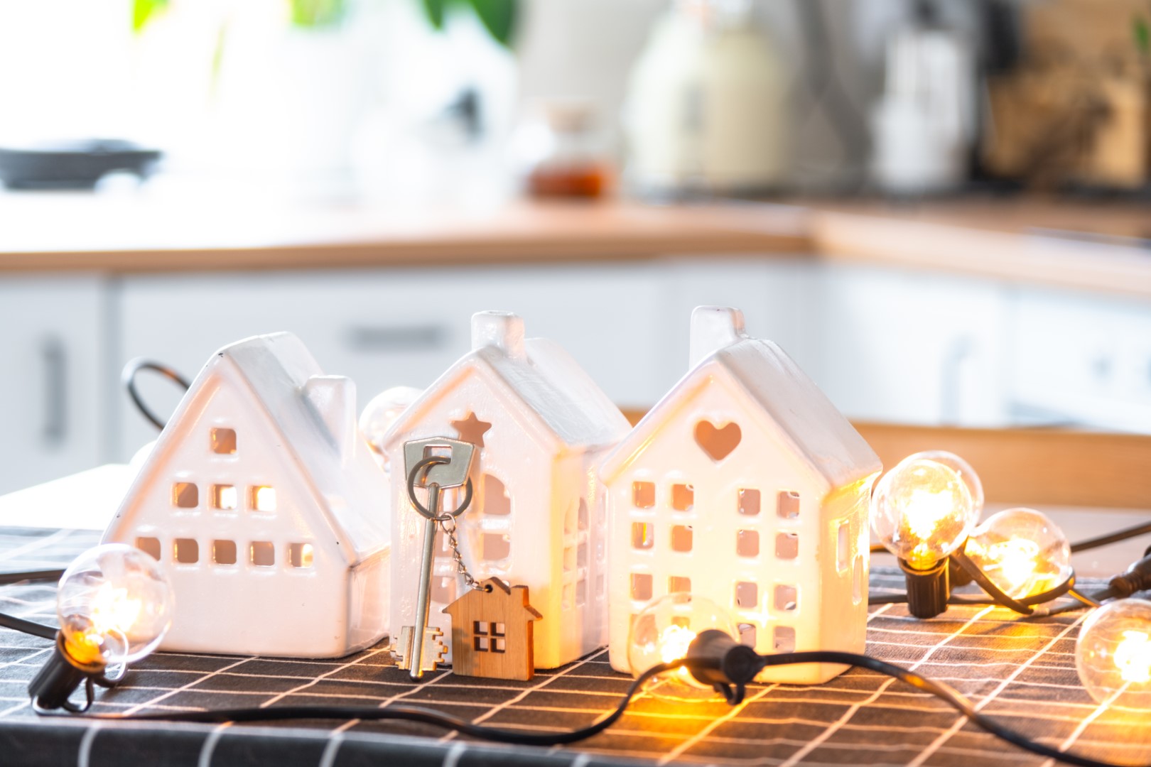 Three white model toy houses, a key, and little bulb lights on a string on a kitchen counter.