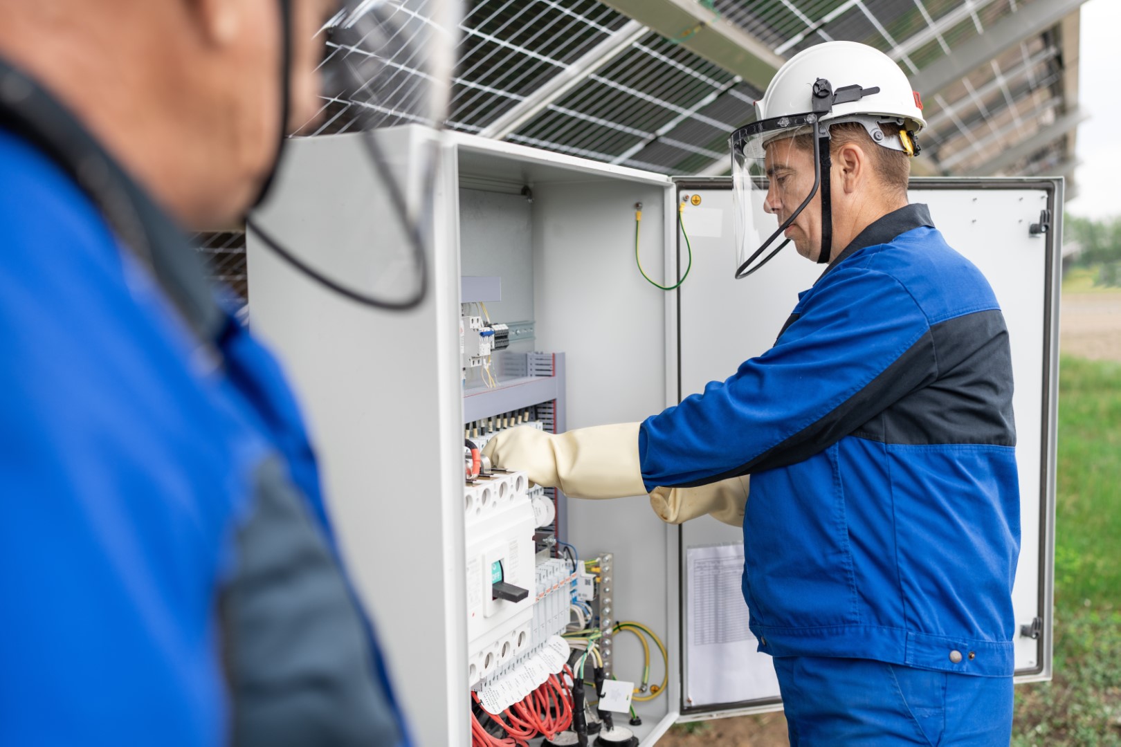 Two electricians in hard hats and blue uniforms working on checking a transformer.