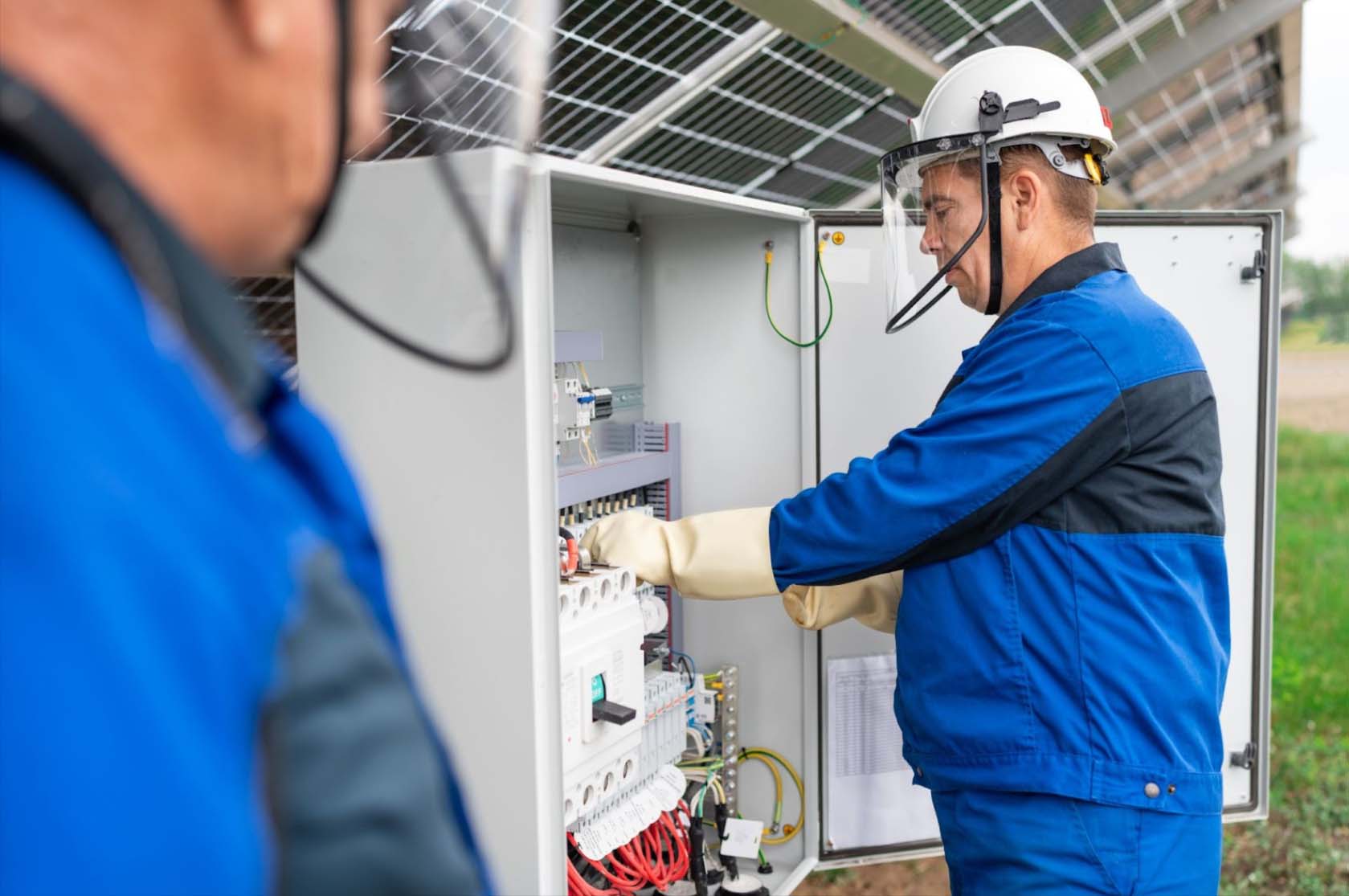 Two male licensed electricians in blue uniforms and hard hats work on checking a transformer site and addressing and solving electrical issues.