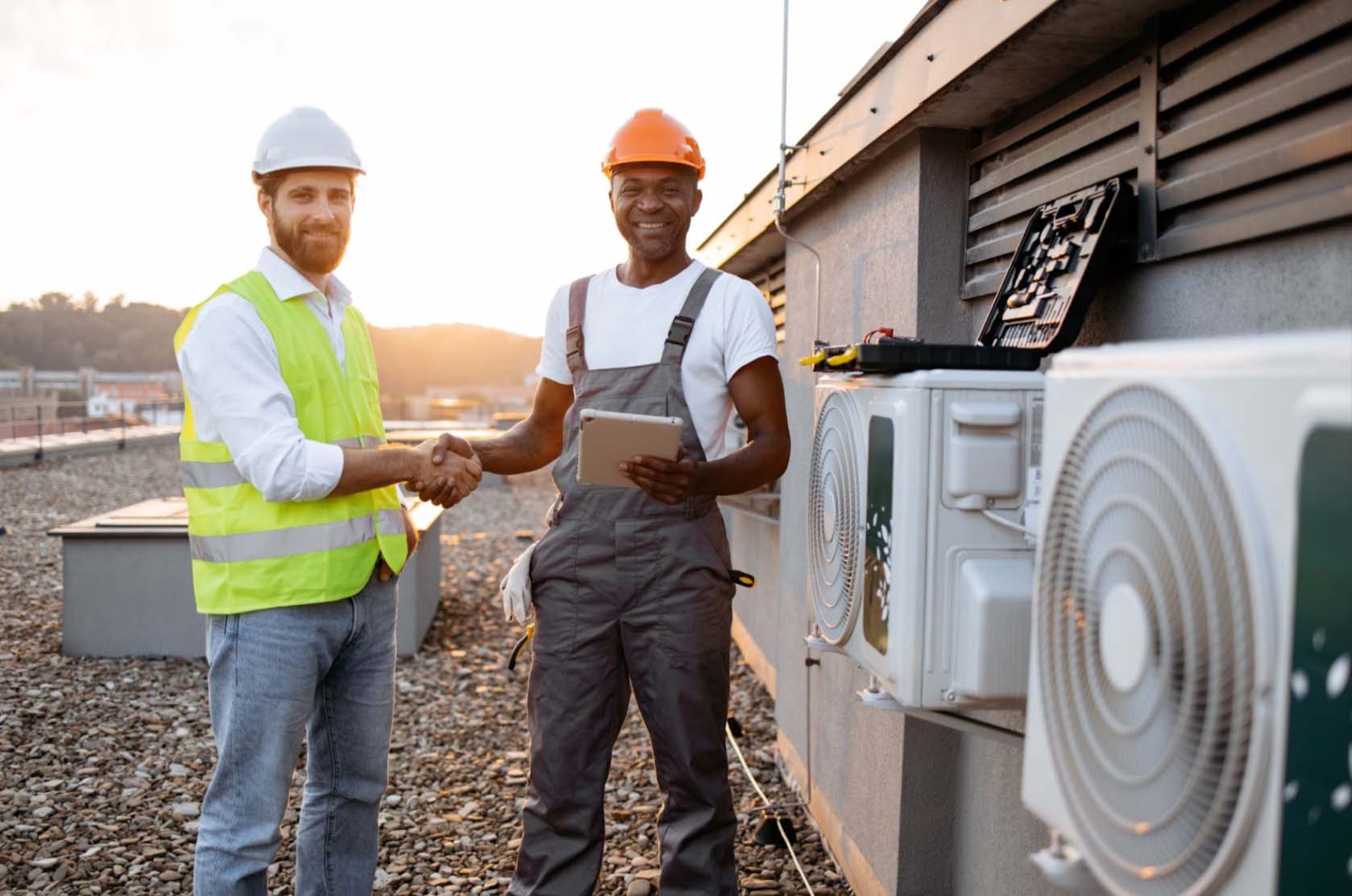 Two skilled electrical contractors shake hands near an outdoor air conditioner after a successful installation. The Caucasian contractor wearing safety gear shakes hands with the African engineer wearing a faded gray jumper, white shirt, and tablet.