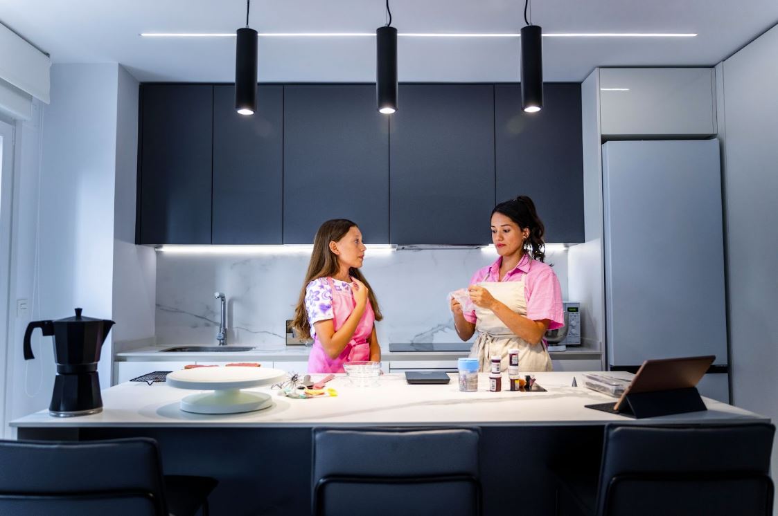 A mother and daughter prepare a pastry bag in a modern gray-themed kitchen with recessed lights and an LED strip light, adding functional, stylish lighting.