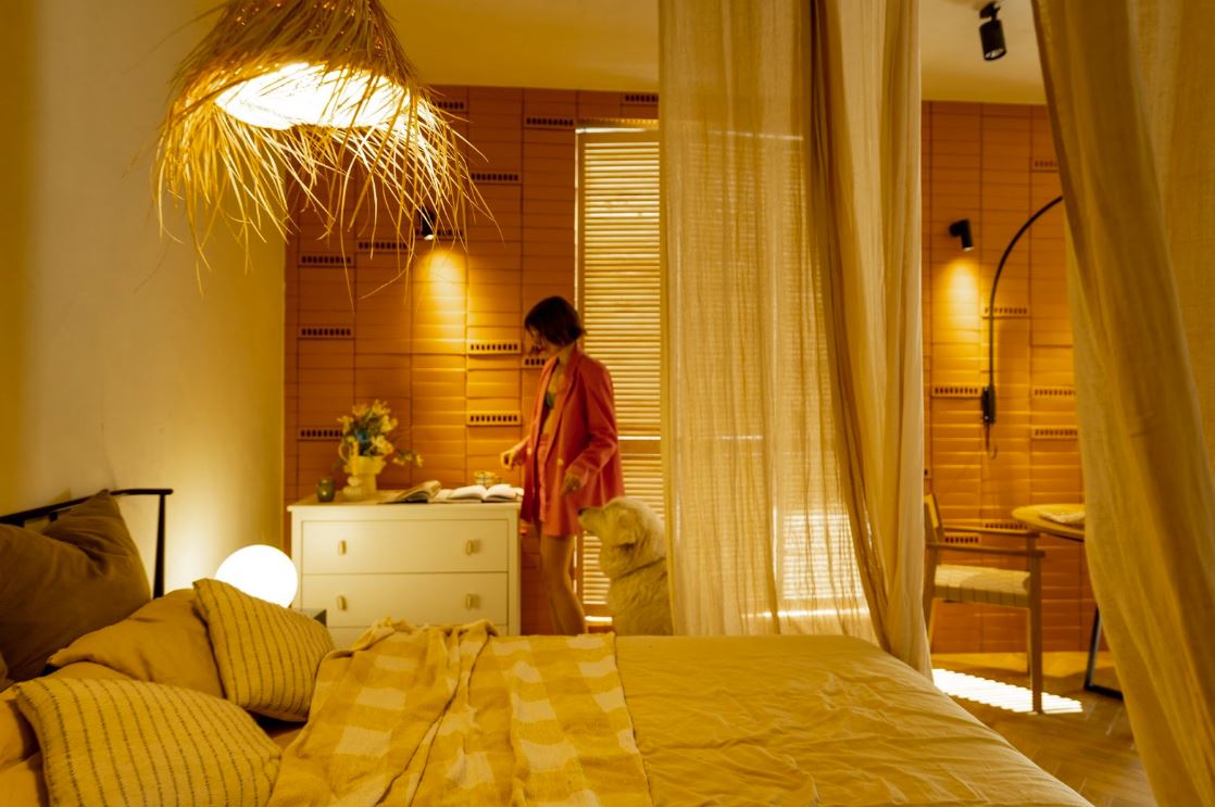 A woman in a red blazer stands by a console table in a beige-toned bedroom featuring a vase of faux sunflowers, a pendant light resembling a beach hat, and black wall sconces.