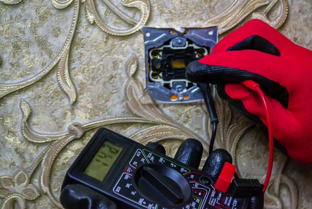 A close-up of an electrician's hand in black and red knitted mitts holding a device while checking a socket on an intricately designed wall.