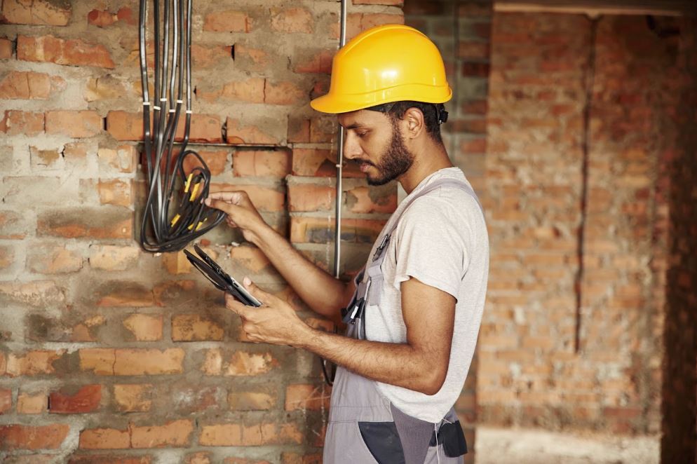 A man holding newly installed wires on brick walls conducts an electrical inspection at a construction site with a digital tablet.