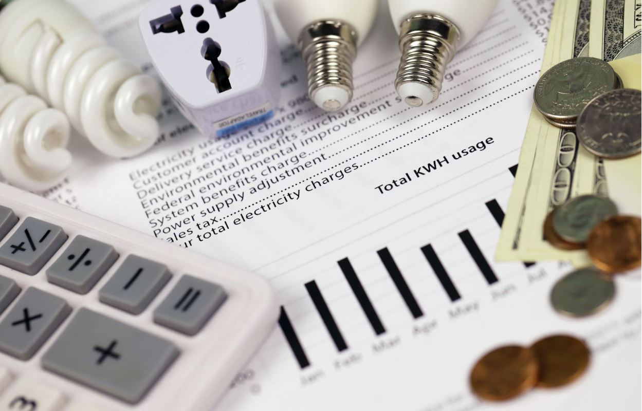 An abstract American electricity bill surrounded by electrical sockets, light bulbs, paper bills, coins, and a calculator.