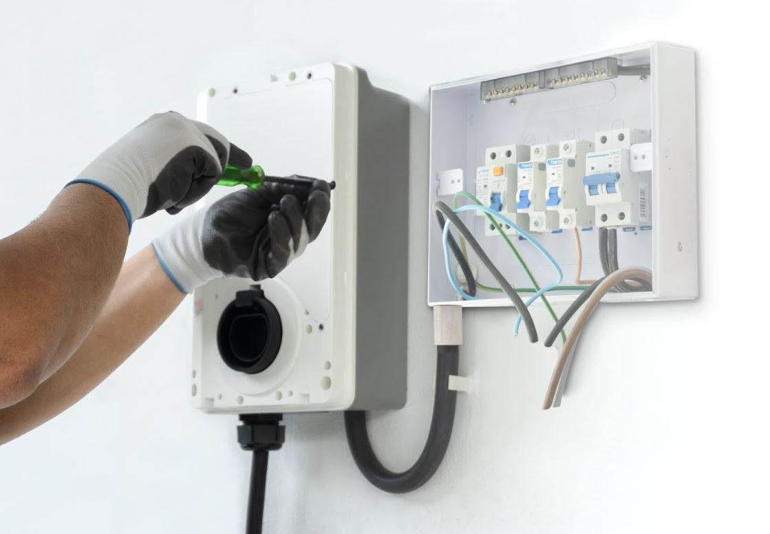 An electrician installs a home EV charging station on a white wall, with only hands and tools visible.