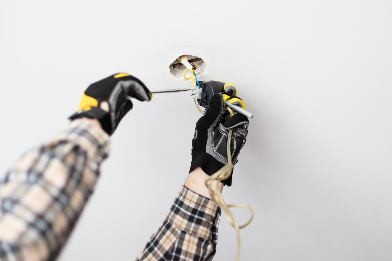 An electrician's arms wearing black and yellow gloves repairing the electrical wiring of a ceiling lamp.