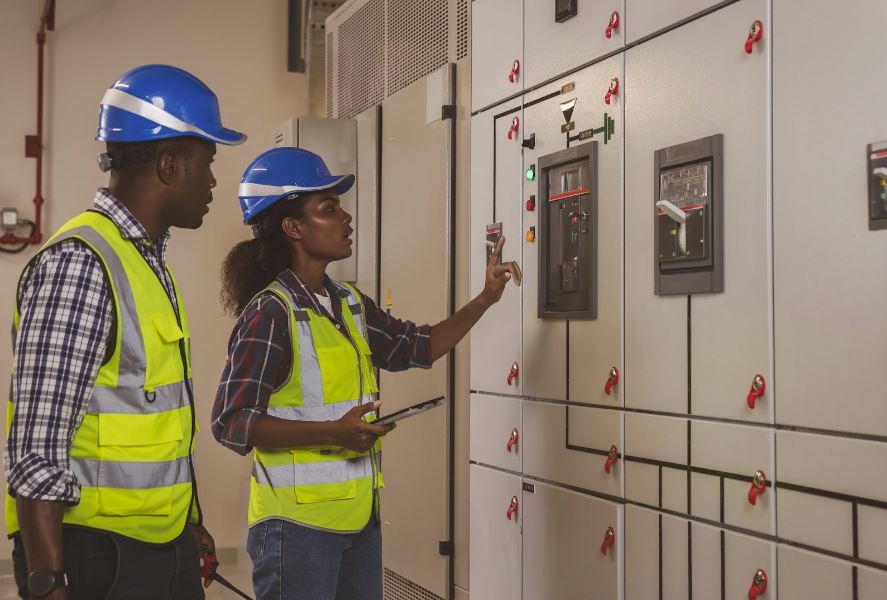 Man and a woman in hard hats and vests looking at an electrical box.